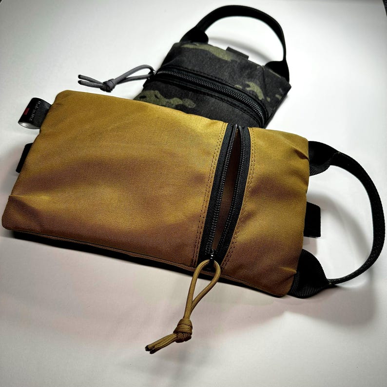 May include: Two rectangular pouches. The front pouch is tan with a black zipper and pull cord. The second pouch is black with a camouflage pattern. Both pouches have a black handle and are on a white surface.
