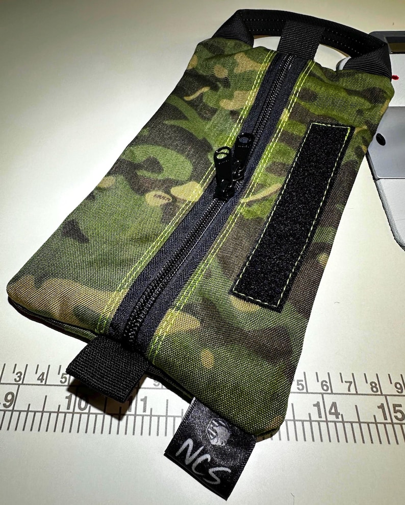 May include: A rectangular camouflage-patterned pouch with a black zipper, handle, and Velcro patch. The pouch has a black label with the letters "NCS" printed on it. The pouch is made of a durable, water-resistant material.