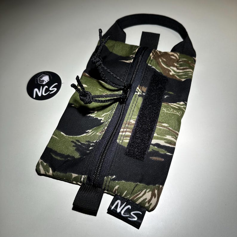 May include: A rectangular pouch with a tiger stripe camouflage pattern in green, black, and brown. It features a black zipper, a Velcro strip, and a black handle. The pouch has the text "NCS" on a small tag and a circular sticker with the text "NCS".