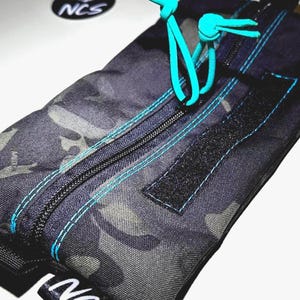 May include: A camouflage-patterned pouch with a black zipper and teal stitching. The pouch has a teal drawstring closure and a black Velcro patch. The logo "NCS" is visible on the pouch and on a circular sticker.