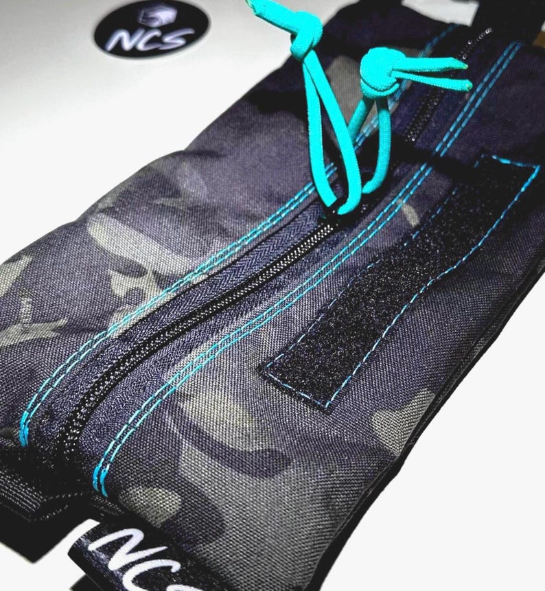 May include: A camouflage-patterned pouch with a black zipper and teal stitching. The pouch has a teal drawstring closure and a black Velcro patch. The logo "NCS" is visible on the pouch and on a circular sticker.