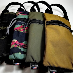 May include: Four vertical pouches with black handles and zippers. The pouches are in various colors and patterns, including black camouflage, teal and pink camouflage, olive green, and tan. Each pouch has a small tag with an American flag.