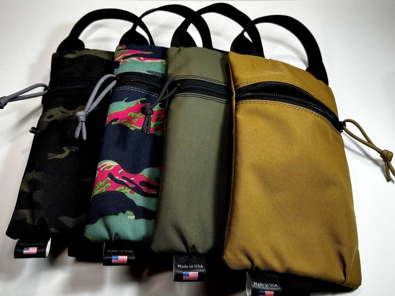 May include: Four vertical pouches with black handles and zippers. The pouches are in various colors and patterns, including black camouflage, teal and pink camouflage, olive green, and tan. Each pouch has a small tag with an American flag.