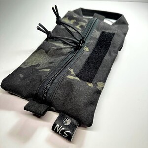 May include: A black camouflage pouch with a zipper closure and a Velcro patch. The pouch has a small tag with the letters "NCS" in white. The pouch is rectangular and appears to be made of durable material.
