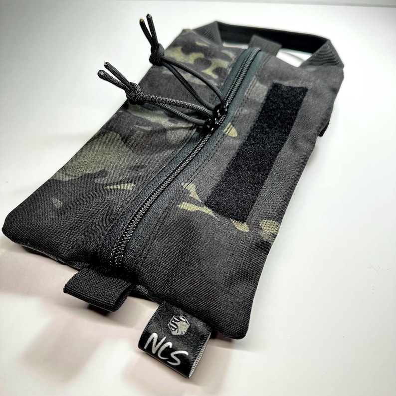 May include: A black camouflage pouch with a zipper closure and a Velcro patch. The pouch has a small tag with the letters "NCS" in white. The pouch is rectangular and appears to be made of durable material.