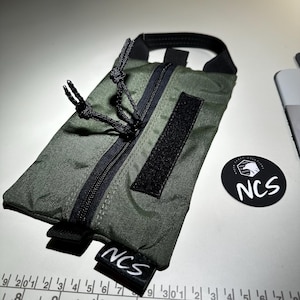 May include: An olive green pouch with a black zipper, handle, and Velcro patch. The pouch has the text "NCS" on a small black tag and a round sticker with the text "NCS". The pouch is approximately 7 inches long.