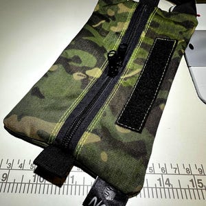 May include: A camouflage pattern pouch with a black zipper, a black Velcro patch, and a black handle. The pouch has a small black tag with the letters "NCS" on it. The pouch is approximately 9 inches long.