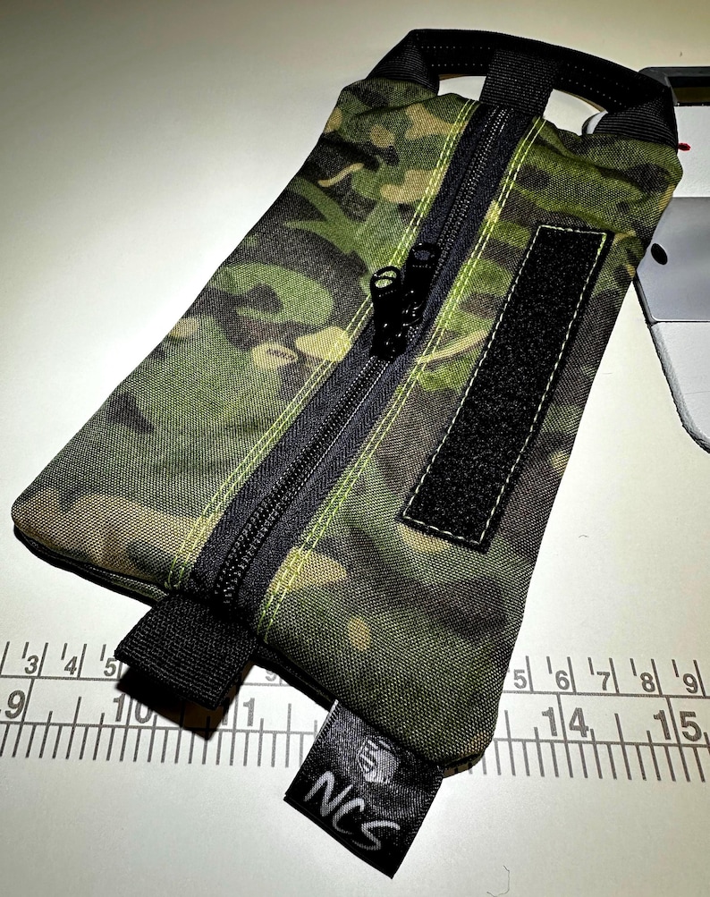 May include: A camouflage pattern pouch with a black zipper, a black Velcro patch, and a black handle. The pouch has a small black tag with the letters "NCS" on it. The pouch is approximately 9 inches long.