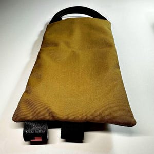 May include: A mustard-colored, trapezoid-shaped bag with a black handle and black straps. The bag has a tag that reads "Made in USA" with a small American flag.