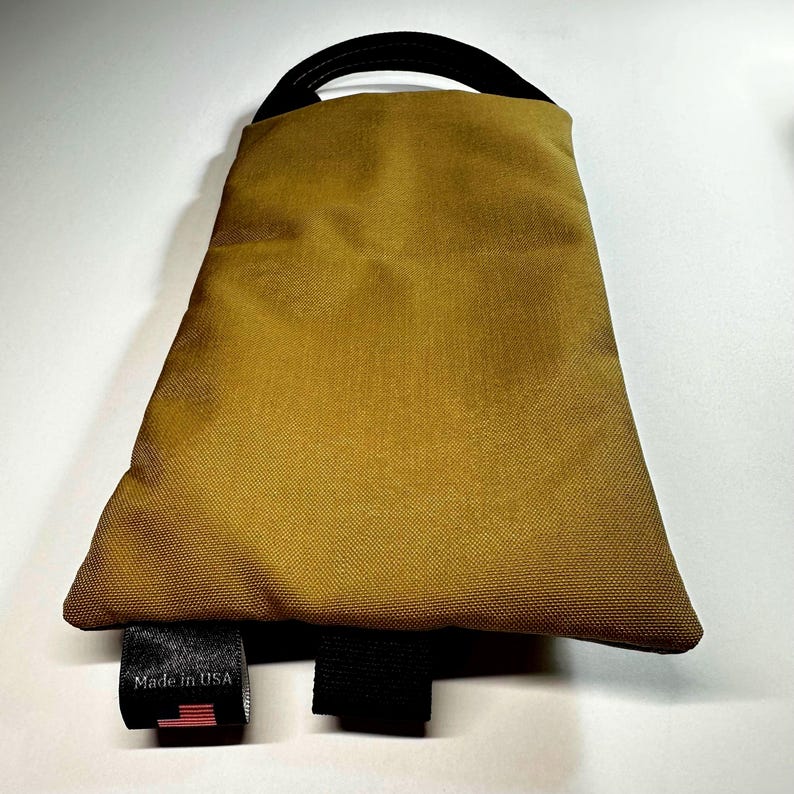 May include: A mustard-colored, trapezoid-shaped bag with a black handle and black straps. The bag has a tag that reads "Made in USA" with a small American flag.