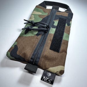 May include: Camouflage pattern pouch with a black zipper, pull cords, and a Velcro patch. The pouch has a black handle and a small tag with the letters "NCS". The color scheme includes shades of green, brown, and black.