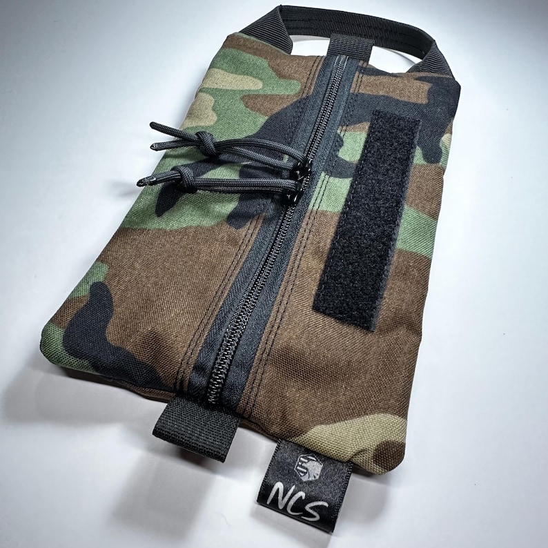 May include: Camouflage pattern pouch with a black zipper, pull cords, and a Velcro patch. The pouch has a black handle and a small tag with the letters "NCS". The color scheme includes shades of green, brown, and black.