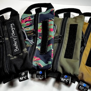 May include: Five zippered pouches in various colors and patterns, including black camouflage, floral, and solid olive and tan. Each pouch has a black handle, a black velcro patch, and a small tag with the text "NCS". One pouch has the text "Battery | Charging".