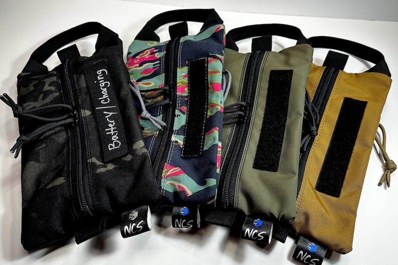 May include: Five zippered pouches in various colors and patterns, including black camouflage, floral, and solid olive and tan. Each pouch has a black handle, a black velcro patch, and a small tag with the text "NCS". One pouch has the text "Battery | Charging".