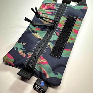 May include: A rectangular pouch with a colorful camouflage pattern in shades of navy, pink, and green. It features a black zipper, a Velcro patch, and a black handle. A small tag with the letters "NCS" is attached.