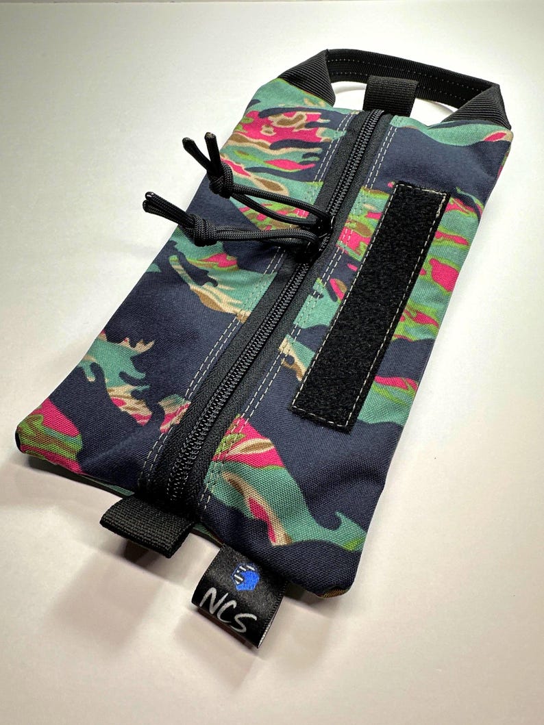 May include: A rectangular pouch with a colorful camouflage pattern in shades of navy, pink, and green. It features a black zipper, a Velcro patch, and a black handle. A small tag with the letters "NCS" is attached.