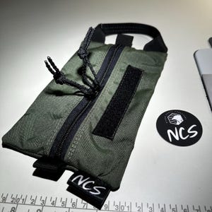 May include: An olive green pouch with a black zipper, handle, and Velcro patch. The pouch has the text "NCS" on a small black tag and a round sticker with the text "NCS". The pouch is approximately 7 inches long.