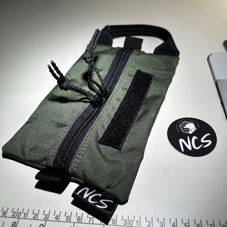 May include: An olive green pouch with a black zipper, handle, and Velcro patch. The pouch has the text "NCS" on a small black tag and a round sticker with the text "NCS". The pouch is approximately 7 inches long.