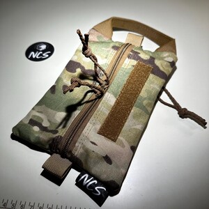 May include: A camouflage pattern pouch with a brown zipper and a loop handle. The pouch has a brown velcro patch and a black tag with the letters "NCS" in white. The pouch is designed for carrying small items.