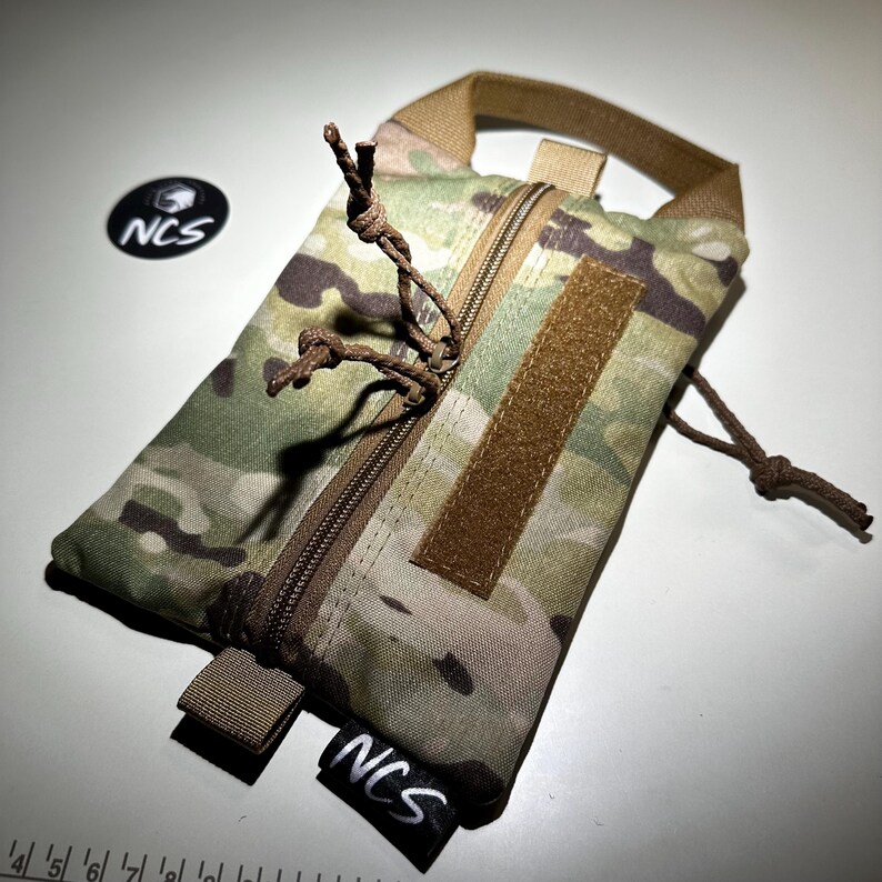 May include: A camouflage pattern pouch with a brown zipper and a loop handle. The pouch has a brown velcro patch and a black tag with the letters "NCS" in white. The pouch is designed for carrying small items.