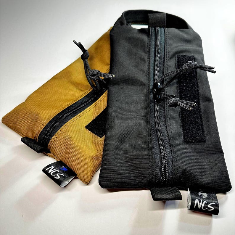 May include: Two rectangular pouches, one mustard yellow and one black, with black zippers and pull cords. Each pouch has a small black tag with the text "NCS" and a logo. The pouches are made of a durable, possibly waterproof material.