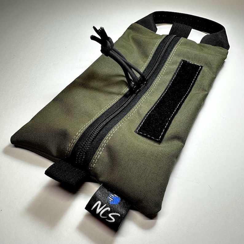 May include: Olive green pouch with a black zipper, pull cord, and a Velcro patch. The pouch has a black handle and a small tag with the text "NCS" and a blue logo. The pouch is rectangular and appears to be made of durable fabric.