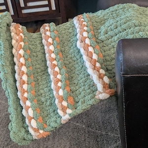 May include: A soft, green knitted blanket with three decorative braided stripes in white, tan, and orange. The blanket is folded over a dark surface, showcasing the texture and color variations.
