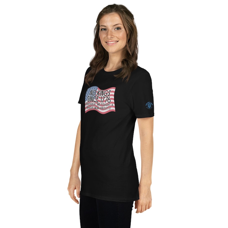 Puede incluir: Camiseta negra con un dise&ntilde;o de bandera estadounidense desgastada y el texto "NO KINGS SINCE 1776". La camiseta tambi&eacute;n tiene un peque&ntilde;o emblema en la manga izquierda. El dise&ntilde;o es en rojo, blanco y azul.