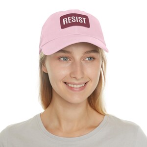 May include: A pink baseball cap with a burgundy patch that says "RESIST" in white letters.