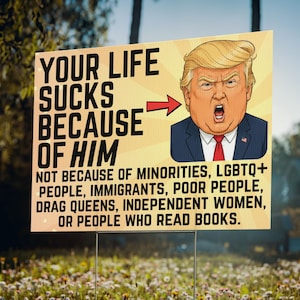 May include: A rectangular yard sign with a yellow background featuring a cartoon illustration of Donald Trump in a blue suit and red tie. The text reads: YOUR LIFE SUCKS BECAUSE OF HIM, with an arrow pointing to the illustration, followed by text.