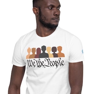 May include: White t-shirt with a graphic of a line of people of different skin tones. The text "We the People" is printed below the graphic in a stylized font.