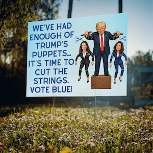 May include: A rectangular political yard sign with a white background and blue text reading WE'VE HAD ENOUGH OF TRUMP'S PUPPETS... IT'S TIME TO CUT THE STRINGS. VOTE BLUE! It features a cartoon of a man standing on a crate controlling two puppets.