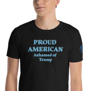 May include: Black t-shirt with the text "PROUD AMERICAN Ashamed of Trump" in blue and white. The shirt has a crew neck and short sleeves. The text is in a bold, sans-serif font.