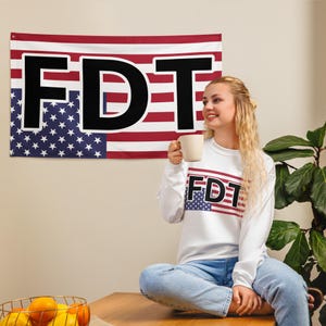 Peut inclure: Un sweat-shirt blanc à manches longues avec le texte "FDT" imprimé en lettres noires. Le texte est sur un petit graphique de drapeau américain. Le sweat-shirt est porté par une personne aux longs cheveux blonds. La personne tient une tasse blanche dans sa main droite.
