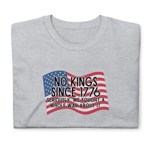 Puede incluir: Una camiseta gris claro con cuello redondo. La camiseta presenta un gr&aacute;fico de una bandera estadounidense ondeando con el texto "NO KINGS SINCE 1776" y la frase "SERIOUSLY, WE FOUGHT A WHOLE WAR ABOUT IT."