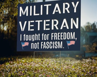 May include: A blue sign with white text that reads "Military Veteran I fought for FREEDOM, not FASCISM." Two small American flags are on either side of the text.