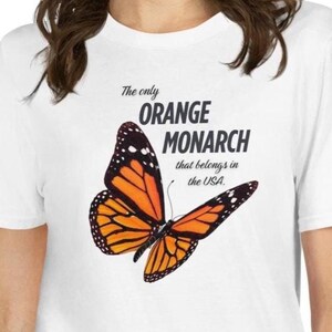May include: White t-shirt featuring a vibrant orange and black monarch butterfly graphic. The text reads "The only ORANGE MONARCH that belongs in the USA." The butterfly design is detailed and eye-catching, perfect for nature lovers.