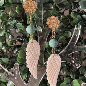 Puede incluir: Un par de pendientes dorados con un diseño floral en la parte superior, una cuenta turquesa y un colgante de ala tallada en blanco roto. Los pendientes se exhiben sobre un fondo de follaje verde.