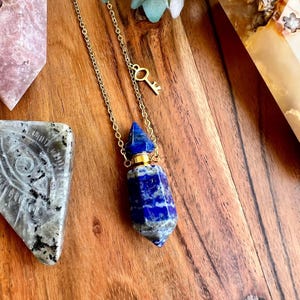 May include: A blue lapis lazuli essential oil bottle pendant with a gold-colored chain and key charm. The gemstone bottle has a faceted design and a gold-colored cap. The necklace is displayed on a wooden surface.