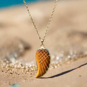 Puede incluir: Un collar dorado con un colgante en forma de ala. El colgante de ala es de color marrón cálido con un patrón texturizado. El collar se muestra sobre una superficie arenosa.