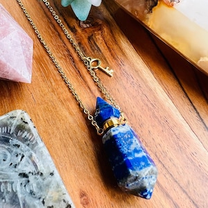 May include: A gold chain necklace featuring a blue and white stone pendant. The pendant is a cylindrical shape with a gold cap and a small key charm. The necklace is displayed on a wooden surface.