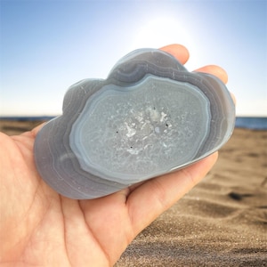 May include: A cloud-shaped, polished agate stone with concentric bands of gray and white. The stone is held in a hand, with a beach and ocean in the background. The agate has a crystalline center and a smooth, polished surface.