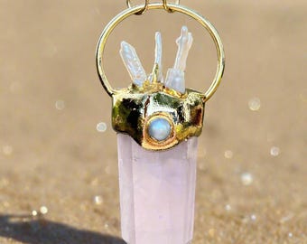 Puede incluir: Un collar dorado con un colgante con una punta de cristal rosa claro. El colgante tiene un anillo dorado y una base dorada con una pequeña piedra redonda azul claro. También hay pequeños cristales transparentes unidos a la base.