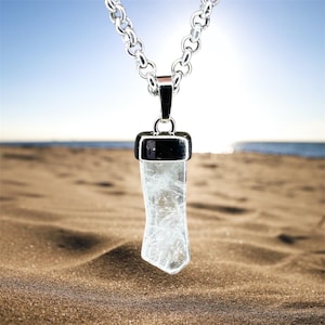 May include: A clear quartz crystal pendant necklace with a silver chain. The crystal is rectangular with a pointed bottom. The necklace is displayed on a sandy beach with the ocean and sky in the background.