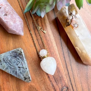 May include: A gold chain necklace featuring a heart-shaped, white stone pendant. The pendant has a small, round top and hangs from a delicate chain. A small key charm is near the clasp. The necklace is displayed with crystals on a wooden surface.