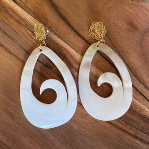 Puede incluir: Un par de pendientes en forma de lágrima con un diseño de ola tallado en la superficie blanca. Los pendientes tienen una parte superior de filigrana dorada. Los pendientes se exhiben sobre una superficie de madera.