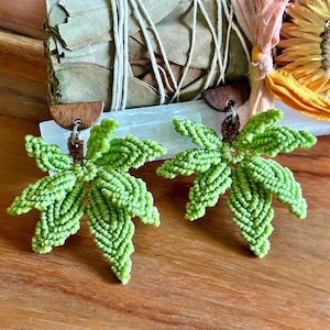 Puede incluir: Un par de pendientes de hojas de macramé verde hechos a mano con detalles de madera. Los pendientes presentan un diseño de hoja detallado, intrincadamente anudado. Se exhiben sobre una superficie de madera, con un manojo de salvia en el fondo.