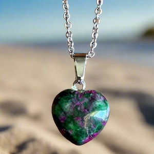 May include: A heart-shaped pendant necklace with a silver chain. The pendant is made of a green and purple stone. The background is a blurred image of a beach.