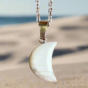Puede incluir: Collar de cadena plateada con un colgante de media luna blanca. El colgante tiene un efecto veteado con tonos grises y una superficie agrietada. El fondo es una playa borrosa.