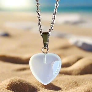 Puede incluir: Collar plateado con un colgante en forma de corazón. El colgante es translúcido, de color blanco roto, y la cadena es de estilo eslabones delicados. El fondo muestra una playa de arena y el mar.